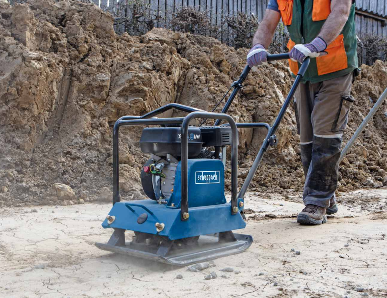 Rüttelplatte auf Baustelle von Person Bedient bedient eine Scheppach-Rüttelplatte auf einer Baustelle. Die Maschine hat einen Motor und wird mit beiden Händen geführt. Im Hintergrund ist ein Erdwall zu sehen.